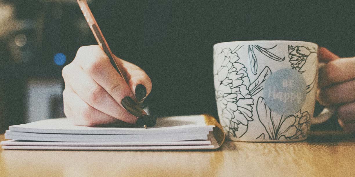 Person writing in a notebook with a cup of coffee on the table