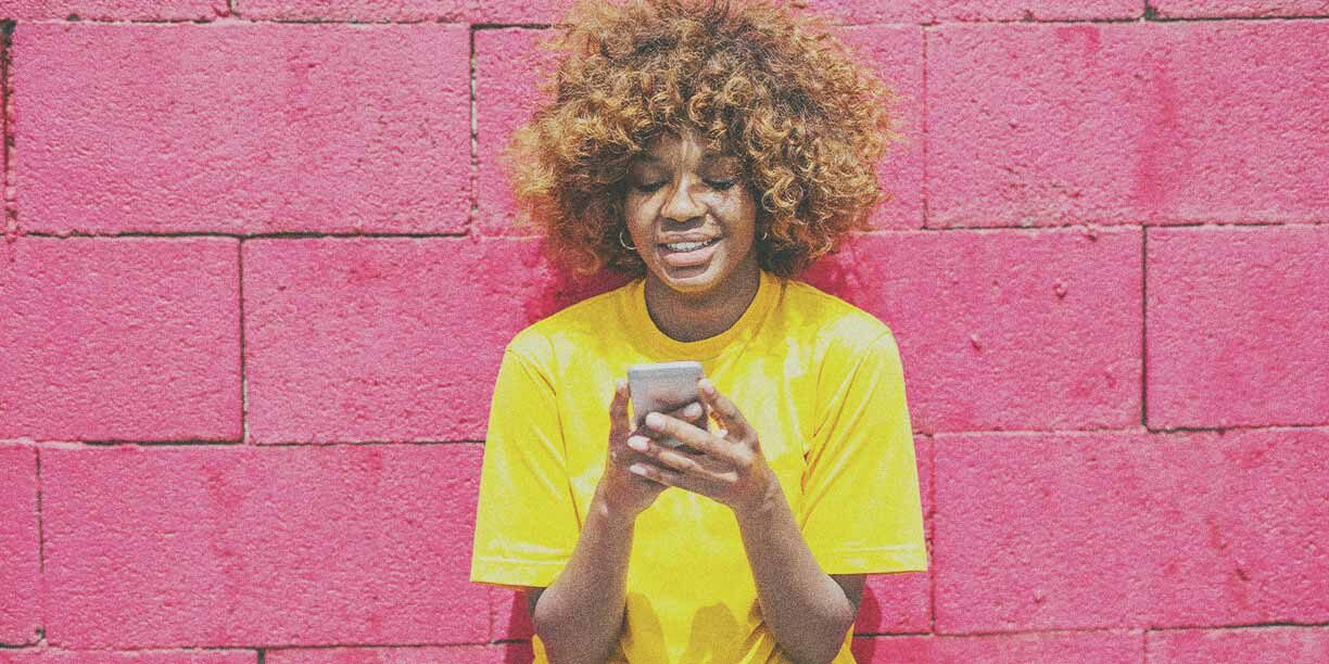 Woman in yellow t-shirt standing against pink wall with cell phone in hand