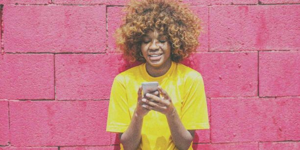Woman in yellow t-shirt standing against pink wall with cell phone in hand