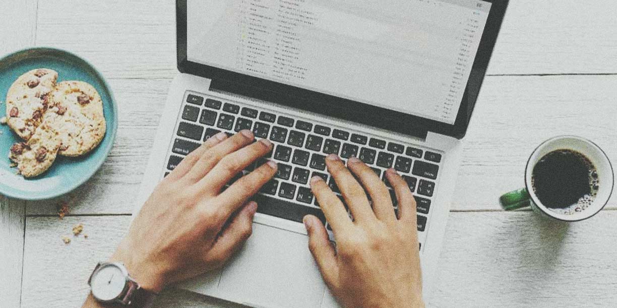 hands typing on laptop with cup of coffee and cookie beside it