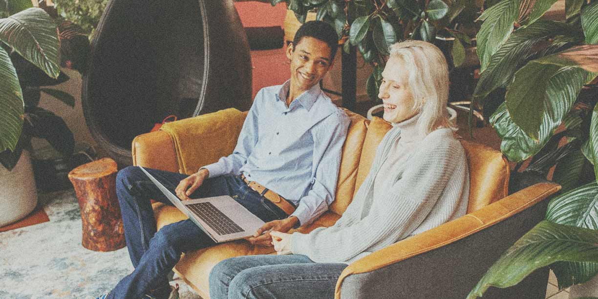 Woman in gray with white hair sitting with a man in blue on a beige leather sofa looking at a shared laptop