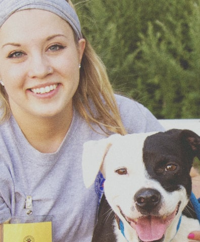 Photo of a young woman wearing a gray tshirt kneeling down and smiling next to a dog
