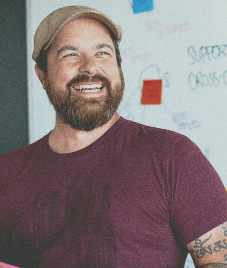 Photo of man with a beard, beige hat and maroon tshirt smiling and looking off camera