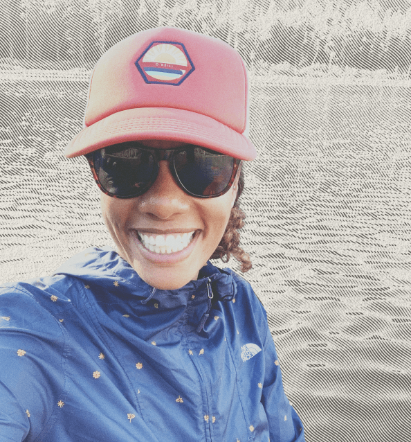 Selfie of smiling woman by a lake in a navy gray jacket, red hat, and sunglasses