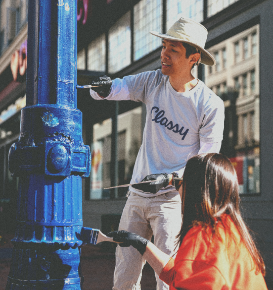 Photo of a man in a gray Classy tshirt and woman in orange sweatshirt outside painting a street lamp