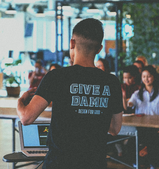 Photo of man from the back in a black tshirt presenting on his laptop to a group of people
