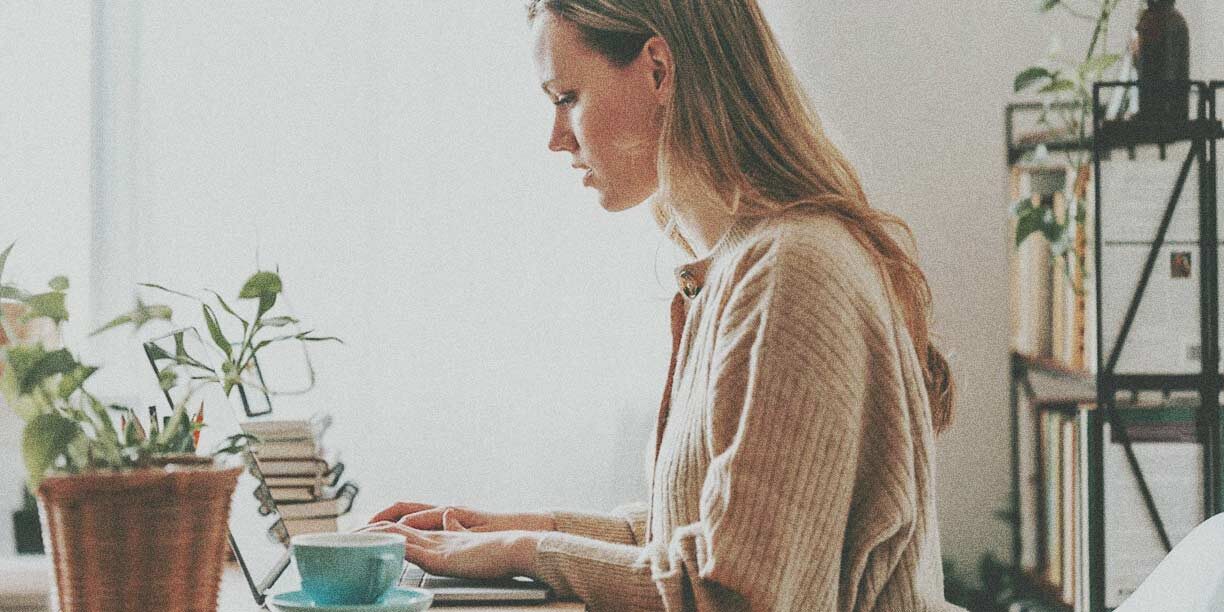 Woman in beige sweater working from home on her laptop at her desk with a cup of coffee