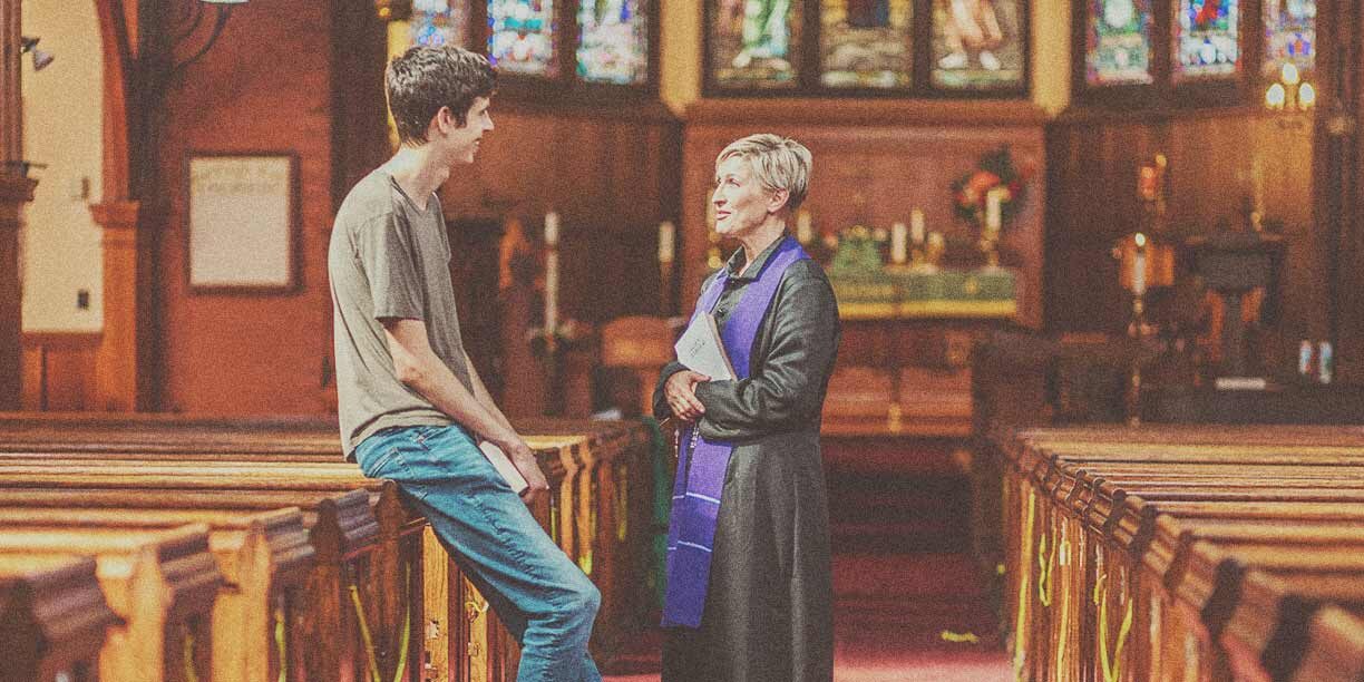 Female pastor standing in a church talking to a young man in a gray shirt and jeans