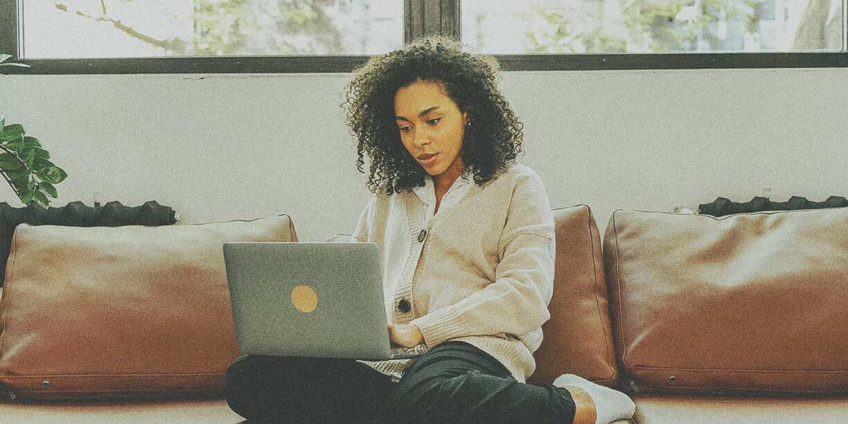 Woman in beige cardigan sitting on a couch typing on a laptop