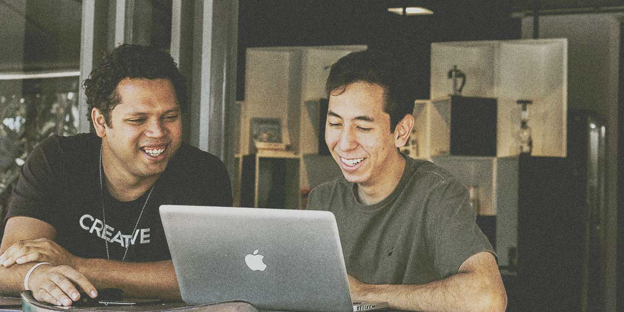 two men in dark shirts sitting at a desk sharing a laptop