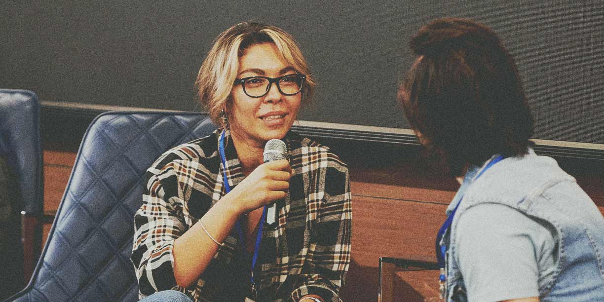 Woman in plaid shirt talking into a microphone to another person in blue