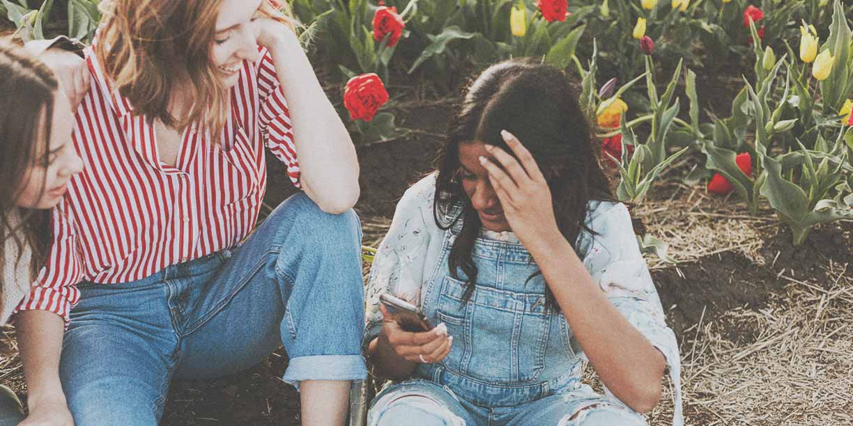 Girl in overalls sitting in a garden with friend in red striped shirt looking at cell phone