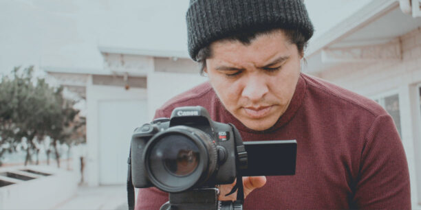 Man in maroon shirt and black hat standing behind a video camera