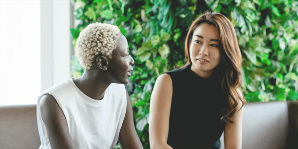 Woman in white talking to a woman in black while sitting on brown leather chairs